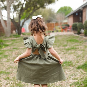 May include: A young girl wearing a green dress with a bow in the back. The dress has ruffled sleeves and a loose fit. She is standing in a grassy area with a house in the background.