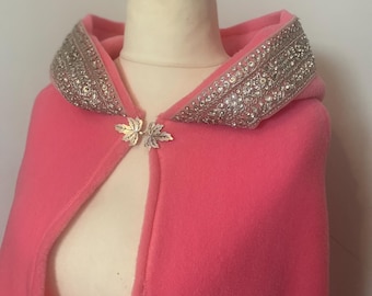 May include: A bright pink hooded cape featuring a wide collar embellished with intricate silver-toned embroidery and sparkling clear crystals. The garment is secured at the neckline with a metallic silver clasp shaped like two joined maple leaves.