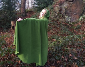 May include: A person wearing a green hooded cloak standing in a forest setting. The cloak is made of a soft, plush fabric and has a long, flowing design.