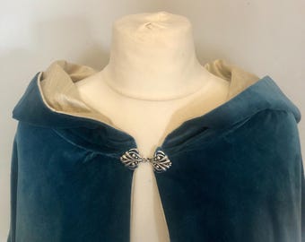 May include: A teal velvet cloak with a hood and a cream-colored lining. The cloak is fastened with an ornate silver clasp. The cloak is displayed on a mannequin.