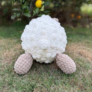 May include: A handmade, plush sheep toy with a fluffy white body and tan legs. The sheep is made of soft, textured yarn and sits on green grass. The toy is designed to be cuddly and is suitable for children.