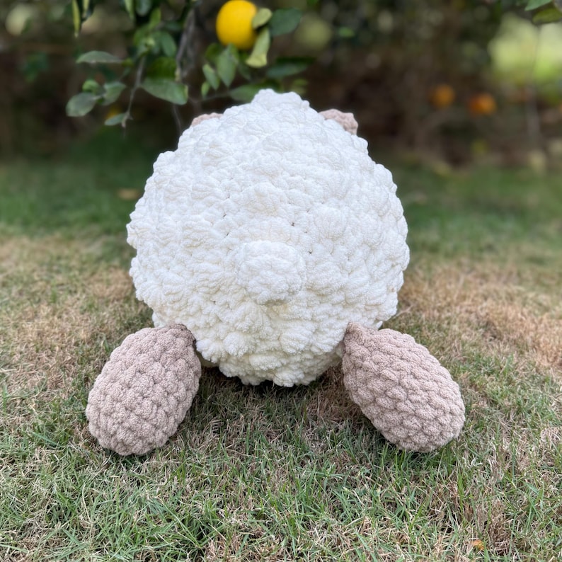 May include: A handmade, plush sheep toy with a fluffy white body and tan legs. The sheep is made of soft, textured yarn and sits on green grass. The toy is designed to be cuddly and is suitable for children.