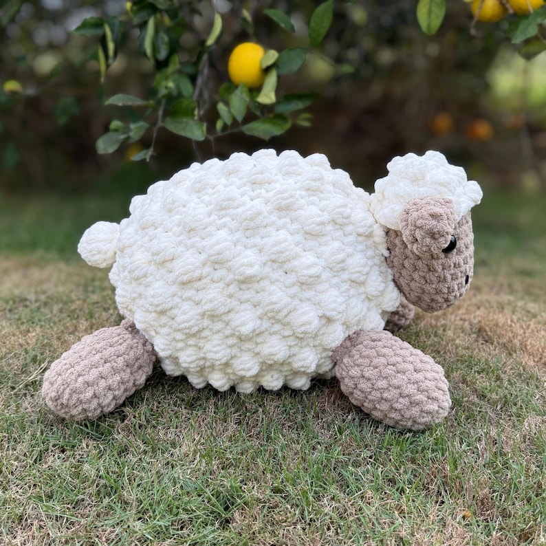May include: A plush sheep toy with a fluffy white body, tan legs, and a tan face with black eyes. The sheep is lying on green grass, with a lemon tree in the background.