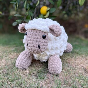 May include: A handmade, plush sheep toy with a fluffy white body and a tan face, ears, and legs. The sheep has black button eyes and is lying on green grass. The toy is designed to look like a cuddly stuffed animal.
