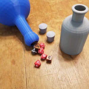 May include: A blue plastic bottle with a wide mouth is tipped over, spilling out a variety of dice. The dice are red, brown, and silver, with white numbers. There are two gray plastic bottle stoppers on the wooden table.