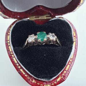 May include: A three-stone ring featuring a central round-cut green emerald flanked by two smaller round-cut clear white diamonds, all set in a metallic band, presented inside a heart-shaped red jewellery box with a black velvet interior lining.