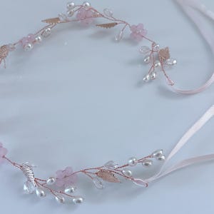 May include: A delicate floral hairpiece featuring pink flowers, faux pearls, and metallic leaves. The headpiece is crafted with a rose gold wire and includes pale pink satin ribbons for securing. A beautiful accessory for weddings or special occasions.