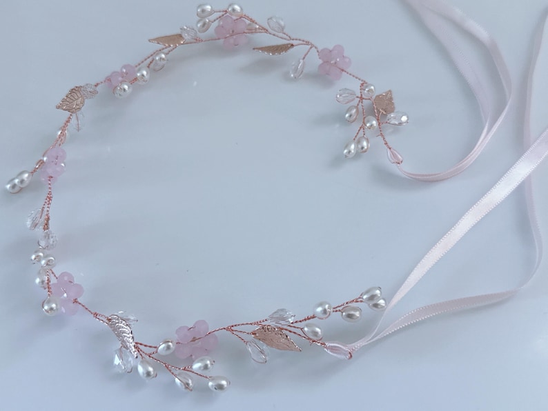 May include: A delicate floral hairpiece featuring pink flowers, faux pearls, and metallic leaves. The headpiece is crafted with a rose gold wire and includes pale pink satin ribbons for securing. A beautiful accessory for weddings or special occasions.
