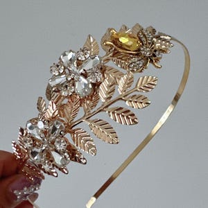 May include: Gold headband with a leaf design and crystal embellishments. The headband features a large crystal flower and a smaller crystal butterfly.