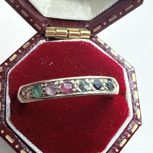 Puede incluir: Un anillo de oro con seis pequeñas piedras preciosas redondas y multicolores dispuestas en fila. Las piedras incluyen tonos verdes, rosas, rojos y azul oscuro. El anillo se presenta dentro de un joyero octogonal forrado de terciopelo rojo.