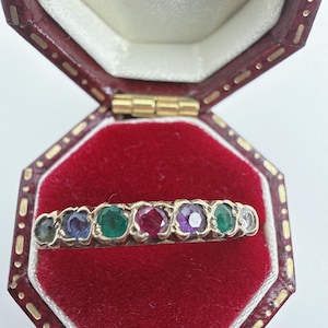 Puede incluir: Un anillo de banda dorada con siete piedras preciosas redondas y multicolores dispuestas en fila. Las piedras incluyen tonos verdes, azules, rojos, morados y blancos transparentes, sujetas por engastes metálicos individuales en la banda.