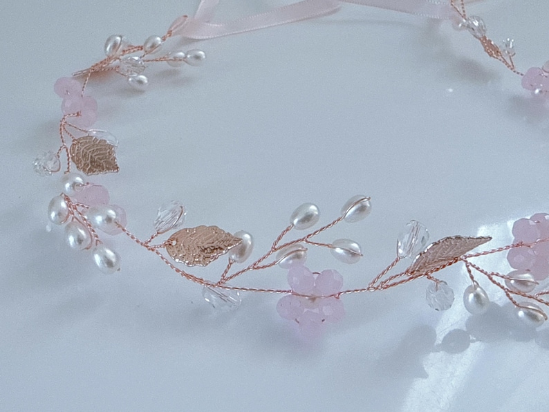 May include: A delicate floral hair accessory featuring a rose gold wire base adorned with faux pearls, pink crystal beads, and metallic gold leaves. A pale pink ribbon is attached for tying. This headpiece is suitable for weddings or special occasions.