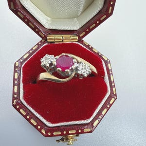May include: A gold ring featuring a central round-cut red ruby flanked by two small round-cut diamonds, presented in an octagonal dark red velvet-lined jewellery box with gold trim and a cream-coloured satin interior lid lining.
