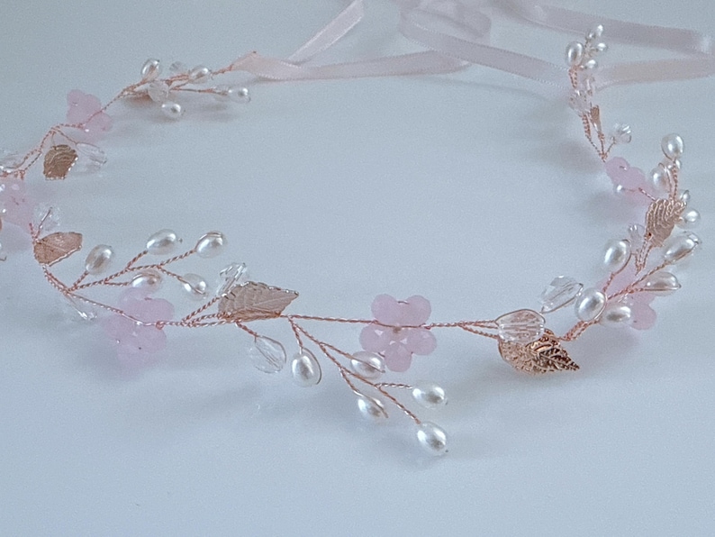May include: A delicate floral headband featuring pink flowers, faux pearls, and rose gold leaves. The wired design includes clear crystal accents and pink ribbons for tying. This elegant headpiece is suitable for weddings or special occasions.