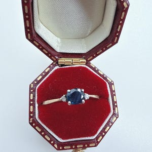 May include: A gold ring featuring a round, dark blue centre stone flanked by two smaller, clear round stones. The ring is displayed inside an open, octagonal, dark red leather jewellery box with a red velvet interior and gold-tone trim.