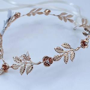 May include: A rose gold-toned metal headband featuring a delicate vine design with sculpted leaves, small rose blossoms, and a butterfly motif. The piece is accented with clear, sparkling crystals embedded within the flowers and leaves, and includes white ribbons.