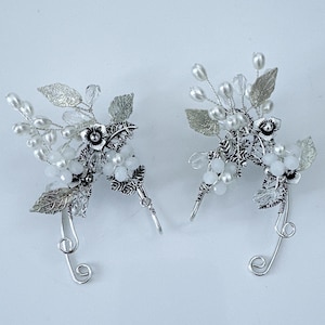 May include: A pair of silver ear cuffs with floral and leaf detailing. Each cuff features silver leaves, white beads, and small flowers. The ear cuffs have a curved design with a wire that loops around the ear.