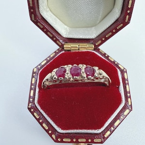 May include: A gold ring featuring three oval-cut red rubies set in a row. The band has an intricate, openwork metal design. The ring is displayed inside an octagonal, dark red leather jewellery box with a cream-coloured interior lining.