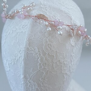 May include: A delicate floral headpiece with a rose gold wire base. The design features small pink flowers, clear crystals, and white pearls. Gold-colored leaves add detail to the piece, which is displayed on a white lace surface.