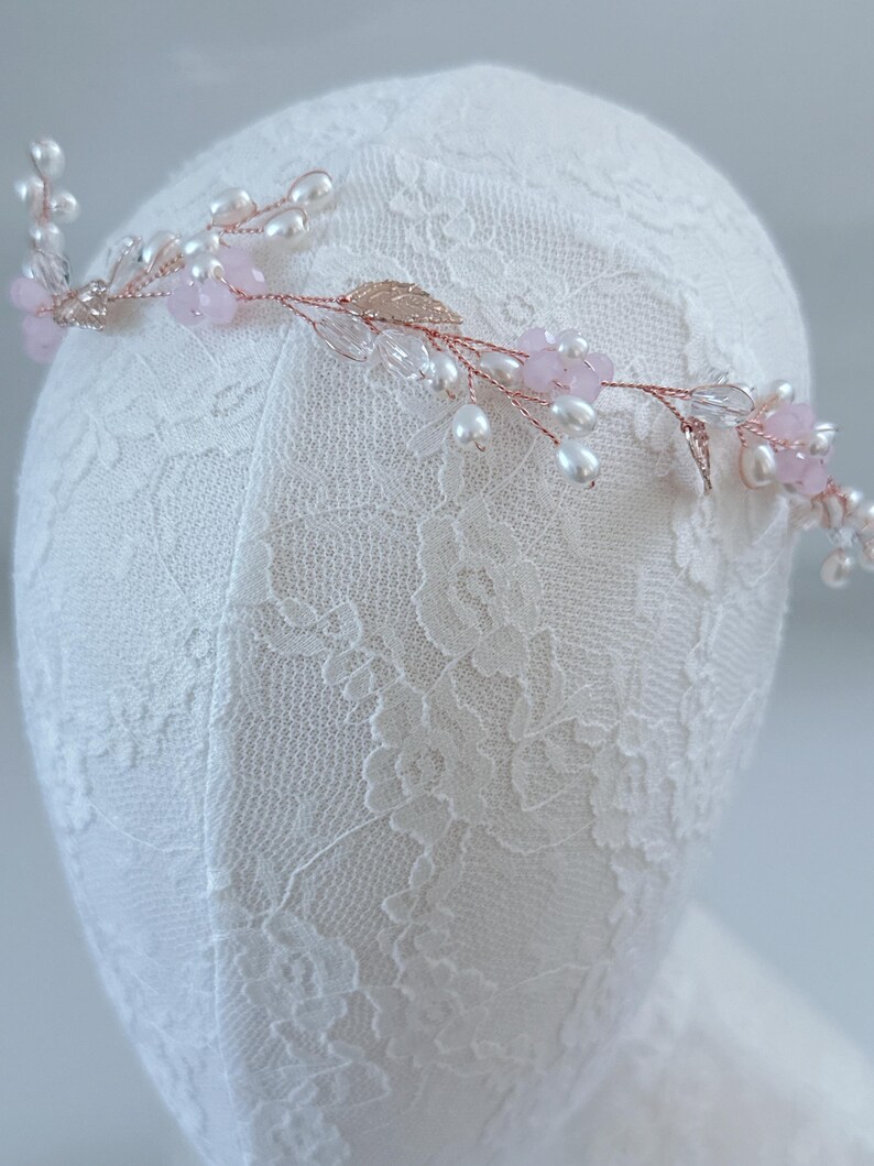 May include: A delicate floral headpiece with a rose gold wire base. The design features small pink flowers, clear crystals, and white pearls. Gold-colored leaves add detail to the piece, which is displayed on a white lace surface.