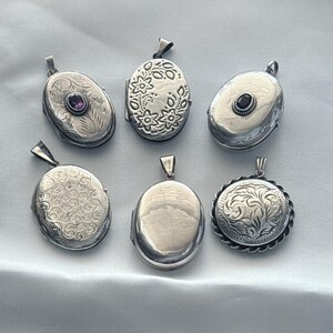 May include: Six silver-toned metal lockets of various shapes and designs. Some feature intricate floral or scroll engravings, while others are plain or set with small purple or dark stones. Each locket includes a metal bail for attachment to a chain.