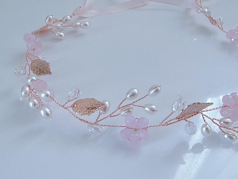 May include: A delicate floral hair accessory featuring a rose gold wire base adorned with pink flowers, faux pearls, clear crystals, and metallic gold leaves. A light pink ribbon is attached for tying. Perfect for weddings or special occasions.