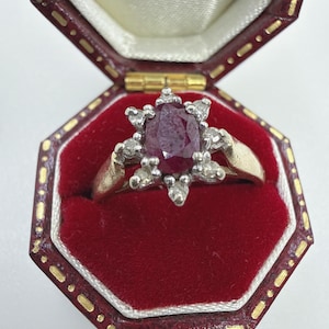 Puede incluir: Un anillo de oro con un rubí rojo intenso de talla ovalada en el centro, rodeado por un grupo de pequeños diamantes transparentes de talla redonda en un engaste de estrella, presentado en un joyero octogonal forrado de terciopelo rojo oscuro.