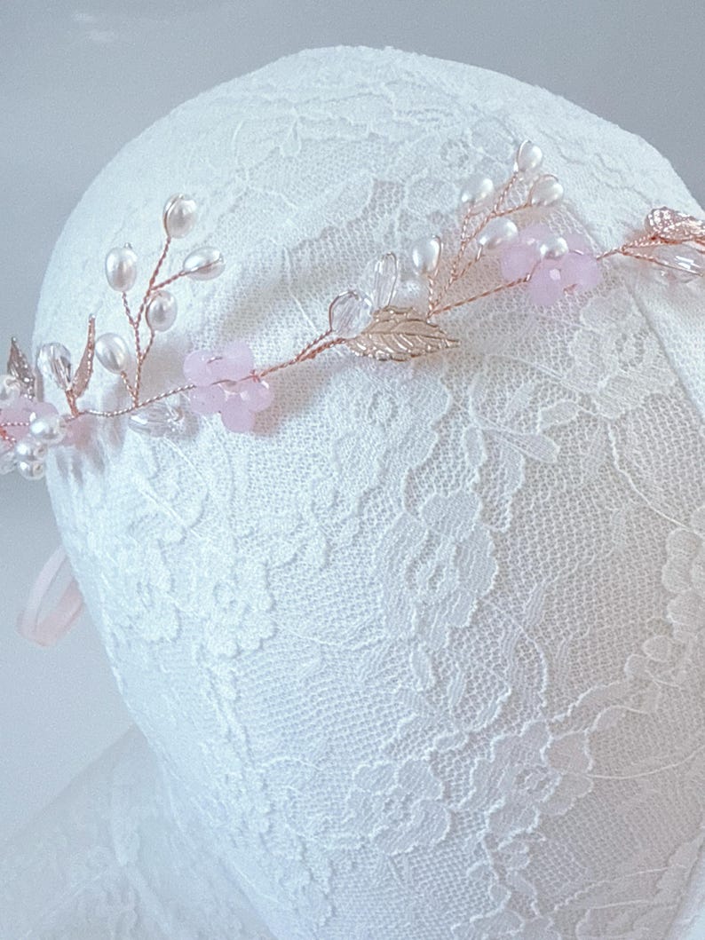 May include: A delicate floral hair crown featuring pink flowers, faux pearls, and gold-colored leaves. The crown is set on a thin, flexible wire, perfect for weddings or special occasions. The design is elegant and romantic.