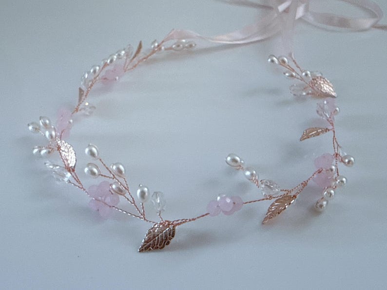 May include: A delicate floral headband featuring a rose gold wire base adorned with faux pearls, pink beads, and metallic leaves. A light pink ribbon is attached for tying. This elegant headpiece is suitable for weddings or special occasions.