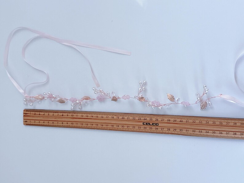 May include: A delicate, pink floral hair accessory with pearl-like beads, rose-colored crystals, and gold-colored leaves. A wooden ruler shows the accessory is approximately 15 inches long. Includes a pink ribbon for tying.