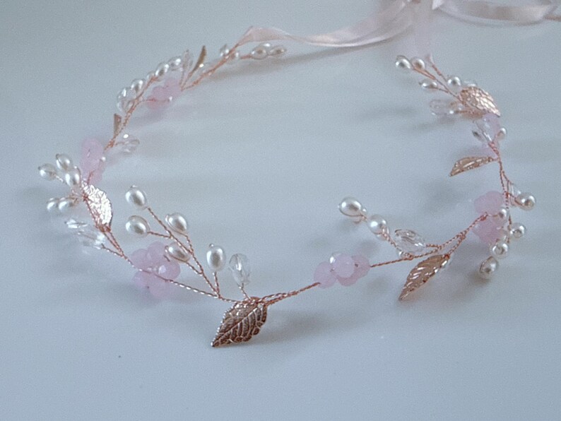 May include: A delicate floral headband with a rose gold wire base. The design features faux pearls, pink flower accents, and metallic gold leaves. A pale pink ribbon is attached for tying.