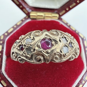 May include: A metal ring featuring an ornate, scrolled surface with a central deep red gemstone flanked by smaller, translucent, light-colored stones. The band displays intricate metalwork patterns across its entire visible exterior surface.