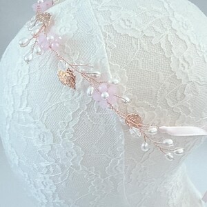May include: A delicate floral headband featuring pink flowers, clear beads, and faux pearls. The headband has gold-colored leaves and is attached to a pink ribbon. The design is set on a white lace background.