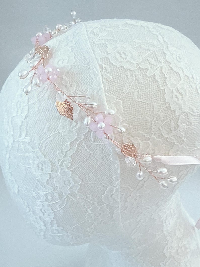 May include: A delicate floral headband featuring pink flowers, clear beads, and faux pearls. The headband has gold-colored leaves and is attached to a pink ribbon. The design is set on a white lace background.