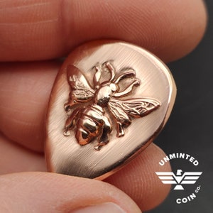 May include: A copper-colored guitar pick with a detailed relief of a bee. The pick has a brushed metal texture and is held between fingers. The logo "UNMINTED COIN CO." is visible.