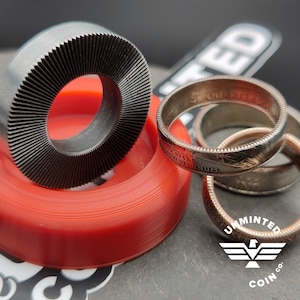 May include: A close-up of a silver coin ring with the words "United States Quarter Dollar" engraved on the band. The ring is on top of a red rubber ring and a black metal ring with a textured surface. The image also includes a sticker with the words "Unminted Coin Co."