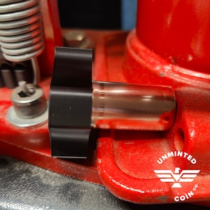 May include: Close-up of a red metal press with a black plastic knob and a copper-coloured cylinder. The press has the text "Unminted Coin Co." printed on the side.