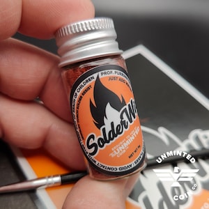 May include: A small glass jar with a silver lid filled with red powder. The label reads "Solder Wire" with a flame graphic and the text "Brought to you by Unminted Coin Co. Net Wt .9g Avoid contact with cap flakes Just add water Prof. Purringtons Jewelry Torch Solder Resist Keep out of reach of children".