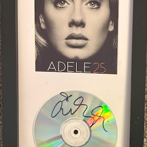 May include: A framed display featuring a black and white portrait of Adele with the text ADELE 25 below it. Below the portrait is a circular compact disc with a holographic surface, featuring a black ink signature written across the center.