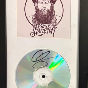 Könnte beinhalten: Ein gerahmtes Display mit einem quadratischen Druck eines bärtigen Mannes mit Cowboyhut und dem Text Chris Stapleton über einer runden Compact Disc. Die Disc hat eine reflektierende, schillernde Oberfläche und trägt eine schwarze Tintensignatur.