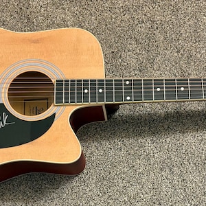 Könnte beinhalten: Akustikgitarre mit natürlicher Holzoberfläche, dunkelroten Zierleisten und einer Signatur auf dem Schlagbrett. Die Gitarre hat ein Schallloch, Saiten und ein Griffbrett mit weißen Einlagen. Die Kopfplatte ist dunkelrot.