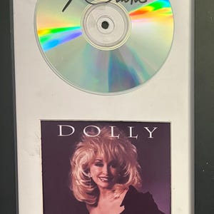 May include: A framed display featuring a signed compact disc with a holographic surface and a black ink autograph. Below the disc is the album cover for Dolly Parton's Something Special, showing a portrait of the artist in a black top.