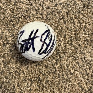 Puede incluir: Una pelota de golf blanca con una superficie con hoyuelos. La pelota tiene firmas manuscritas de color azul oscuro en su superficie. La pelota descansa sobre una superficie marrón y texturizada.
