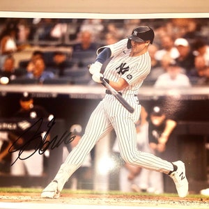 May include: A photograph of a baseball player in a New York Yankees uniform, mid-swing. The player is wearing a pinstriped uniform with the number 18. The image is signed. The background shows other players and spectators.