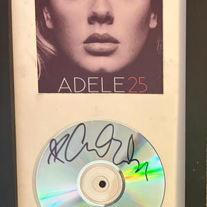 Könnte beinhalten: Ein gerahmtes Display mit dem Adele 25 Albumcover, das ein Porträt der Künstlerin über dem Text ADELE 25 zeigt. Unter dem Albumcover befindet sich eine kreisförmige Compact Disc mit einer schwarzen Tintensignatur auf der reflektierenden Oberfläche.
