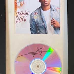 Può includere: CD e copertina dell'album incorniciati con Jimmie Allen. La copertina dell'album mostra il testo "Mercury Lane" e "Jimmie Allen". Il CD è firmato con un pennarello nero. La cornice è nera con un passe-partout bianco.