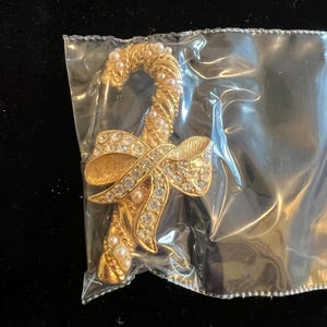 May include: A gold-toned candy cane brooch with a bow, encrusted with clear rhinestones and faux pearls. The brooch is in a clear plastic bag, set against a black background. The candy cane shape is a classic holiday design.