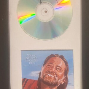 May include: Framed CD and album art featuring Willie Nelson. The CD is signed in black marker. The album art below features a portrait of Willie Nelson with the text "Willie Nelson's Greatest Hits (A Song That Will Be)" in white.
