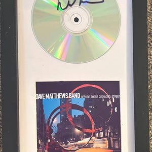 May include: A framed display featuring a signed compact disc and the album cover art for Dave Matthews Band Before These Crowded Streets. The CD has a black ink signature on its reflective surface, and the square album art shows a city street.