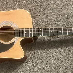 Puede incluir: Una guitarra acústica de madera natural con una golpeador negro y un acabado rojo-marrón en la pala. La guitarra tiene una forma de cuerpo con un corte y un diapasón de palisandro.